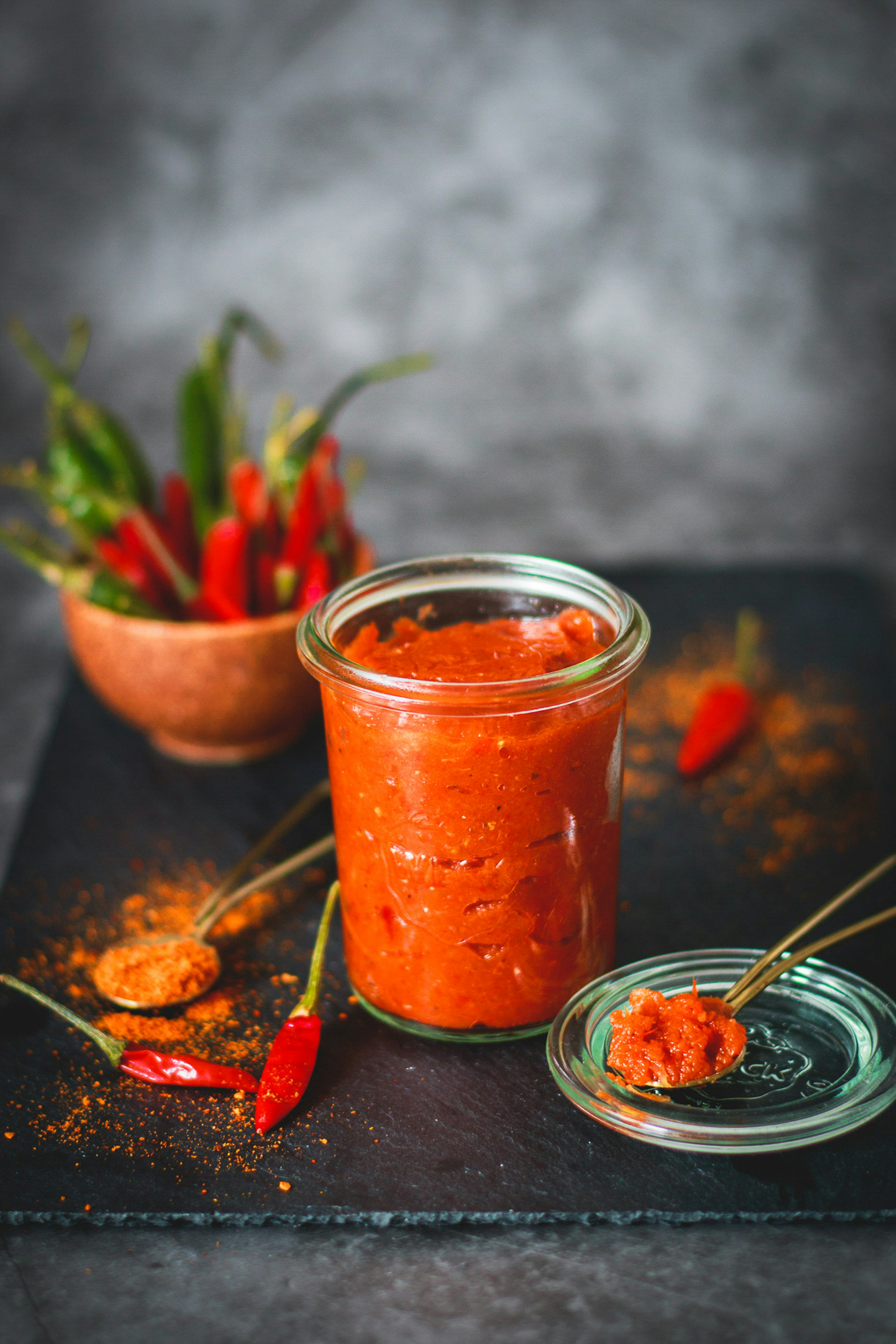 Homemade chili sauce in a glass jar with fresh red chillies and spices on a dark slate board